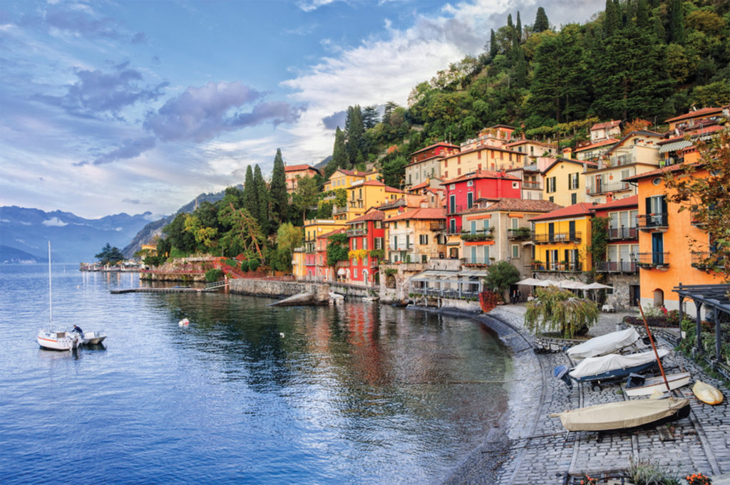 Red and yellow Italian style buildings at water's edge, wooded mountain behind, small boats on sloping cobbled foreshore