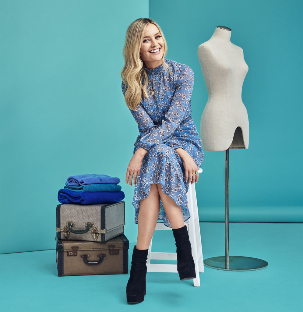 Young blonde woman in black ankle boots and high necked, long sleeved blue floral dress, sitting on stool, turquoise background