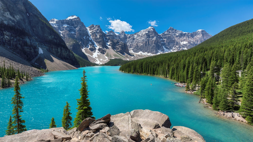 Bright turquoise lake, snow-dusted mountains beyond, forest and rocky shore