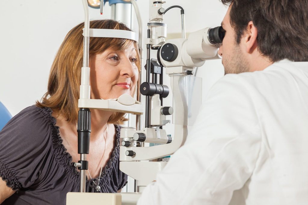 Ophthalmologist In Exam Room With Mature Woman Sitting In Chair Looking Into Eye Test Machine; 