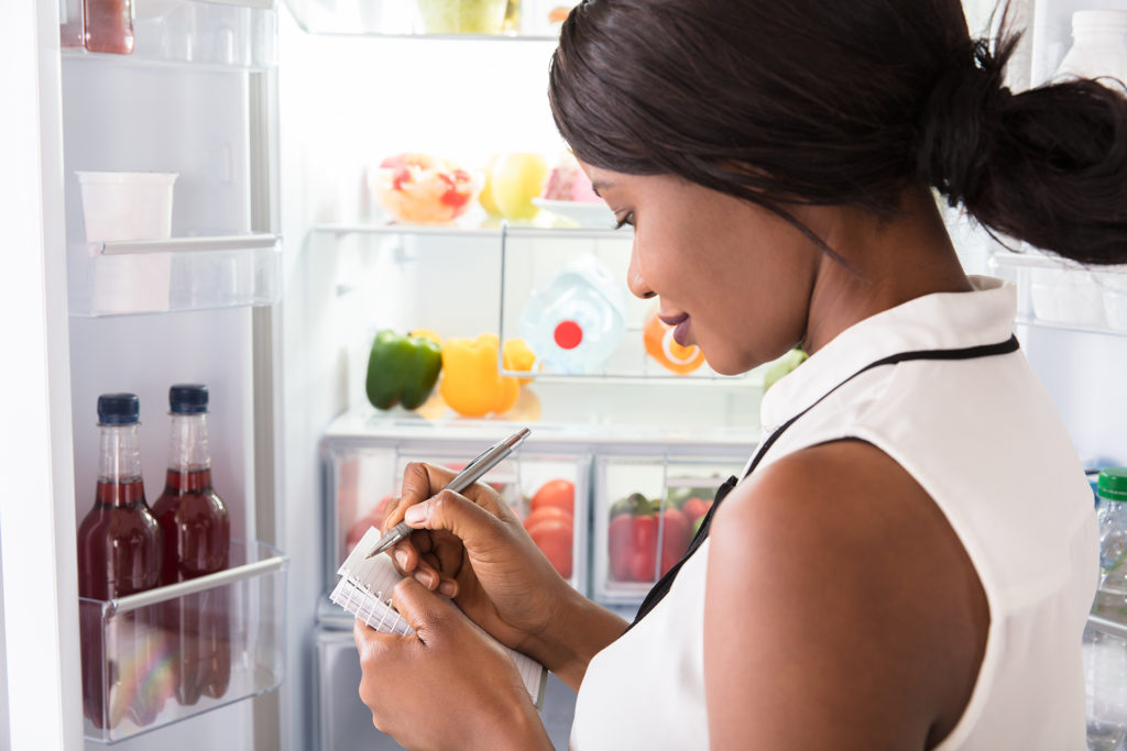 Close-up Of Young African Woman Writing On Spiral Book Near Open Refrigerator; 