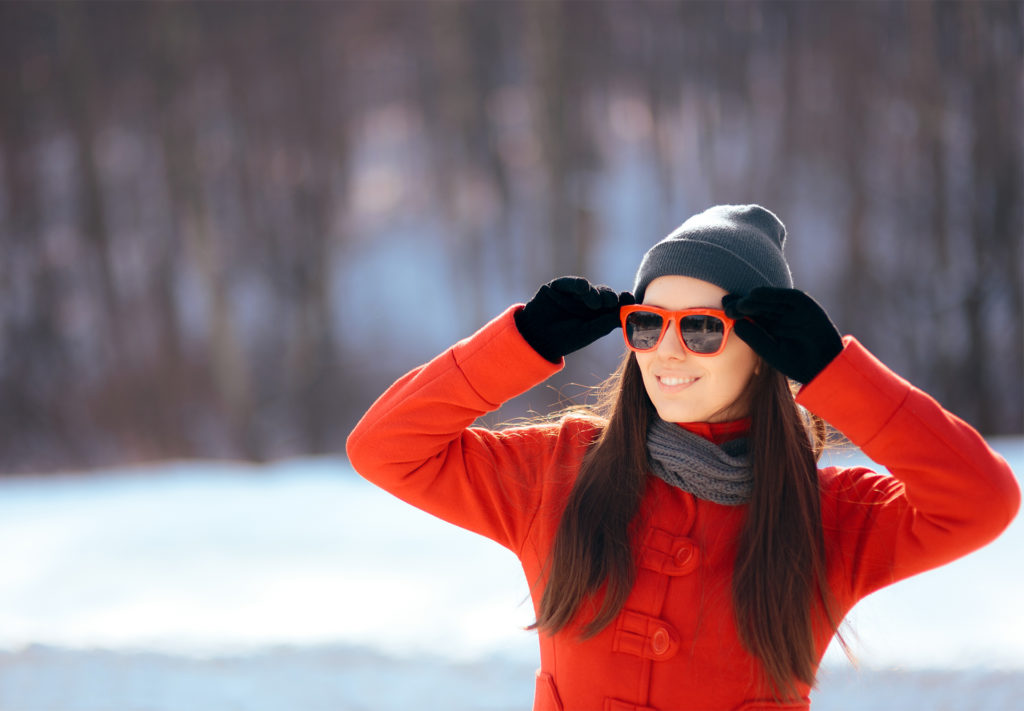 Woman in hat, scarf, red jacket and red framed sunglasses in snow
