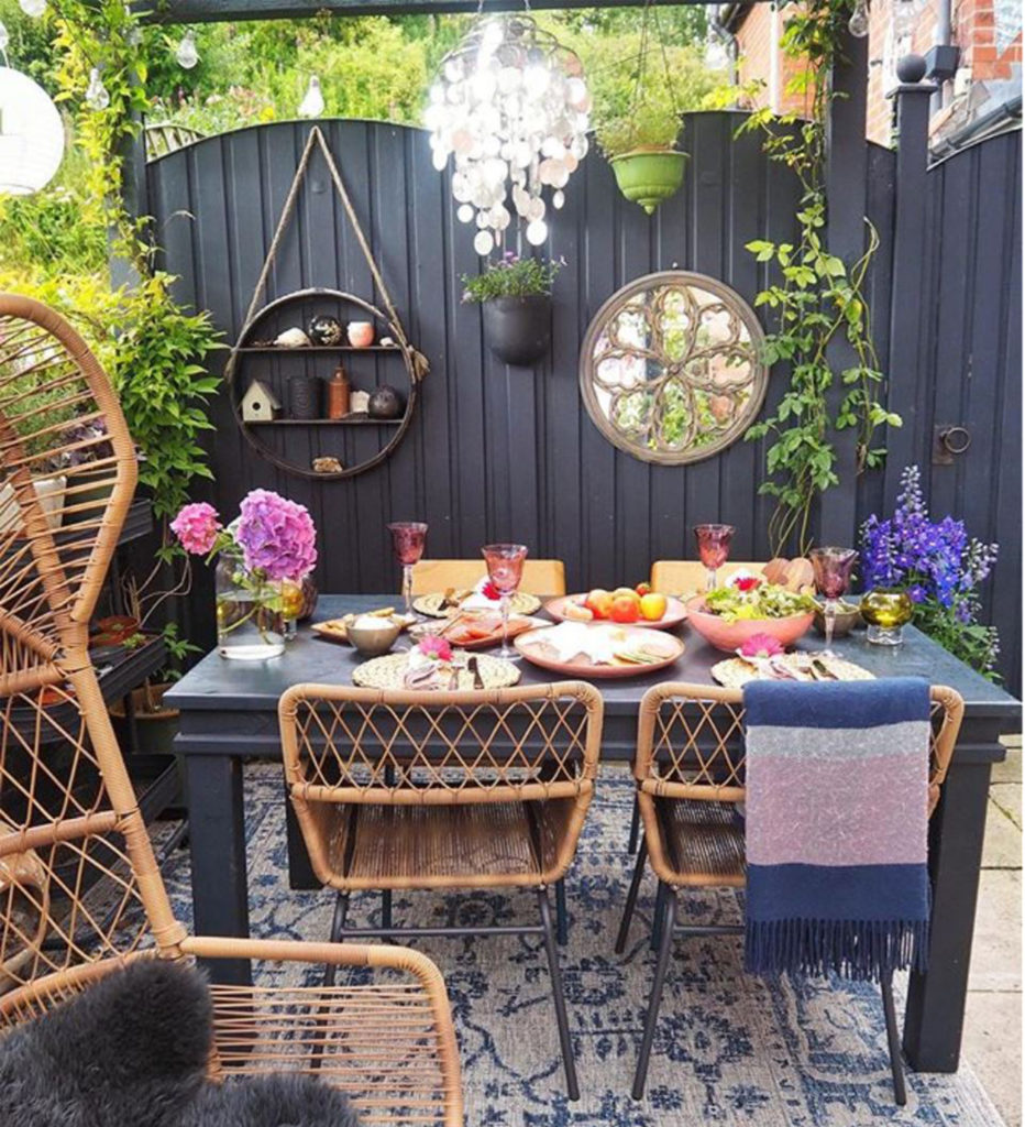 Table and chairs on patio, high grey fence with mirror, climbing plants and decorative shelves