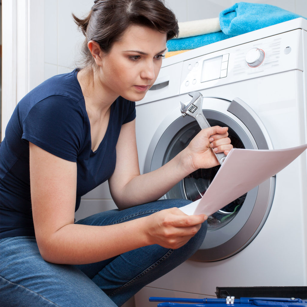 A woman holding a spanner kneels by washing machine and reads instructions