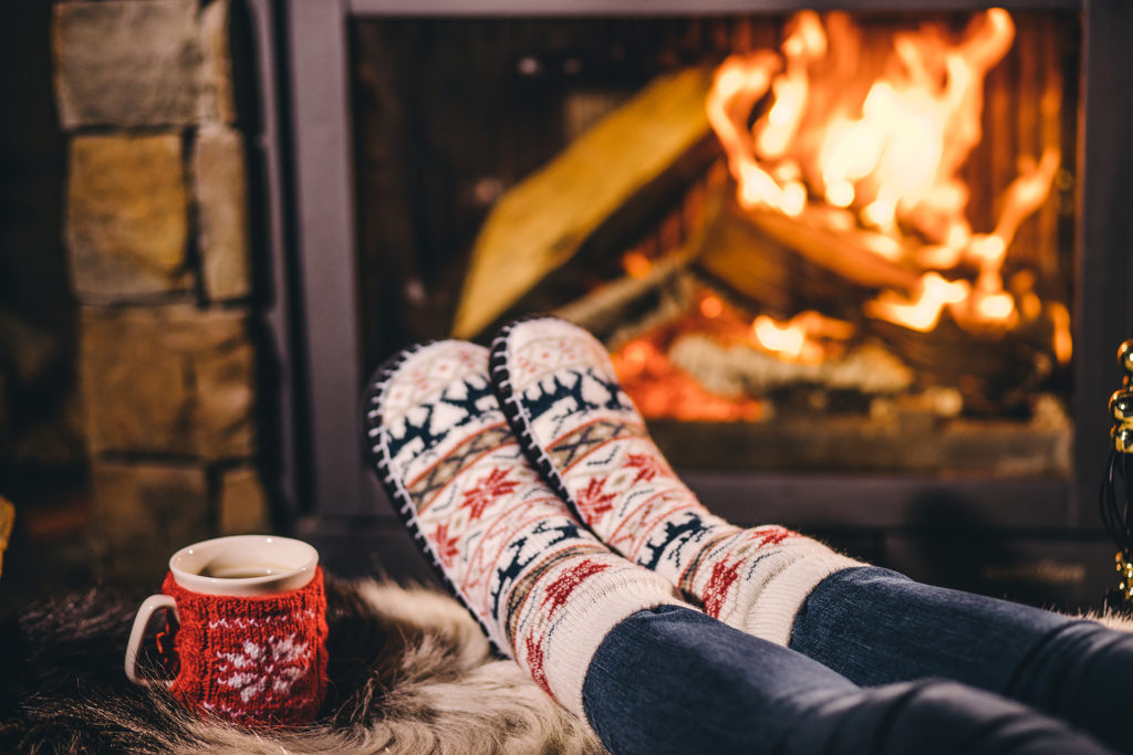 Feet in woollen socks by the Christmas fireplace. Woman relaxes by warm fire with a cup of hot drink and warming up her feet in woollen socks. Close up. Winter and Christmas holidays concept.