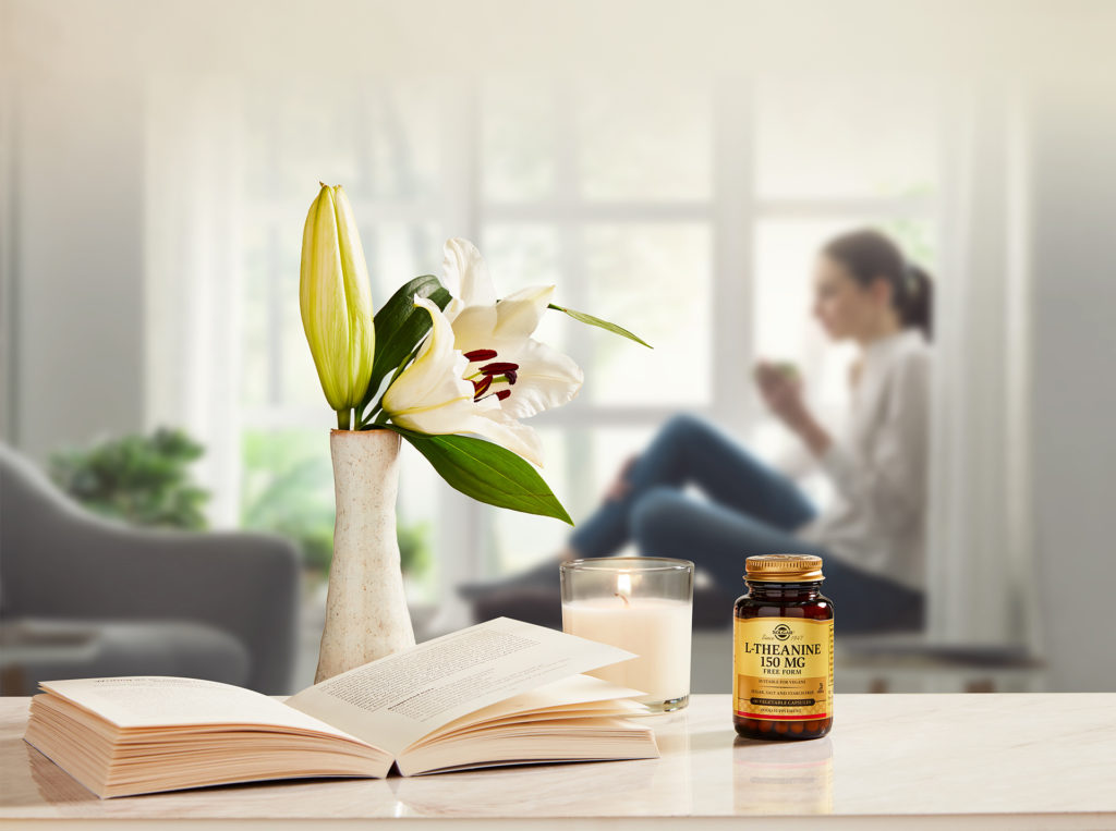 Woman sits by window with cup. In foreground are a vase of lilies, an open book and a bottle of Solgar vitamins