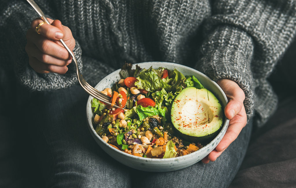 Healthy vegetarian dinner. Woman in jeans and warm sweater holding bowl with fresh salad, avocado, grains, beans, roasted vegetables, close-up. Superfood, clean eating, vegan, dieting food concept; 