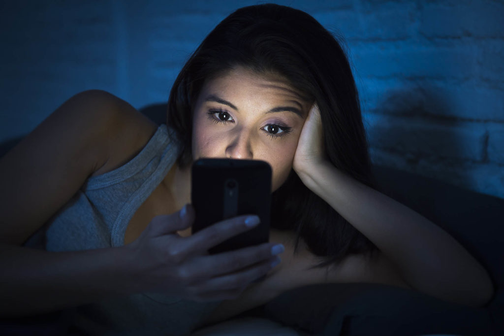 young beautiful woman in bed using mobile phone late at night at dark bedroom lying happy and relaxed enjoying social media network at her phone in communication internet addiction concept; 