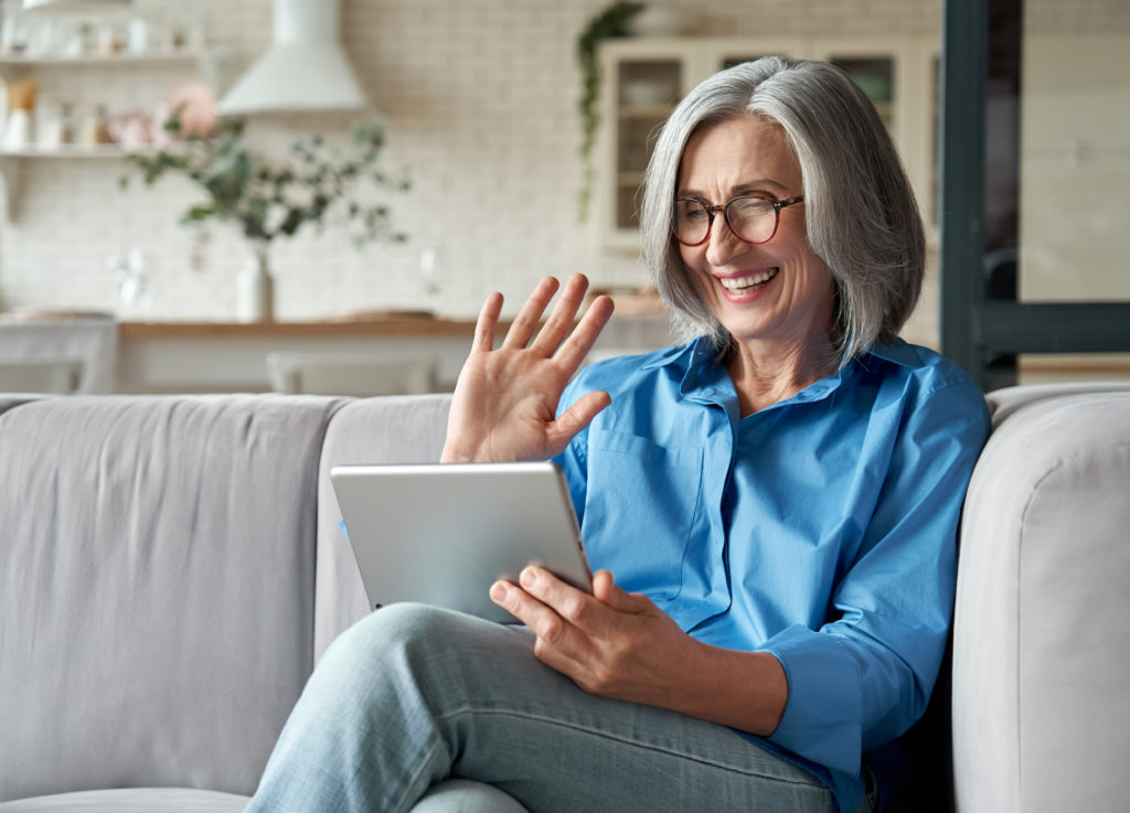 Happy 60s older mature middle aged adult woman waving hand holding digital tablet computer video conference calling by social distance virtual family online chat meeting sitting on couch at home.;