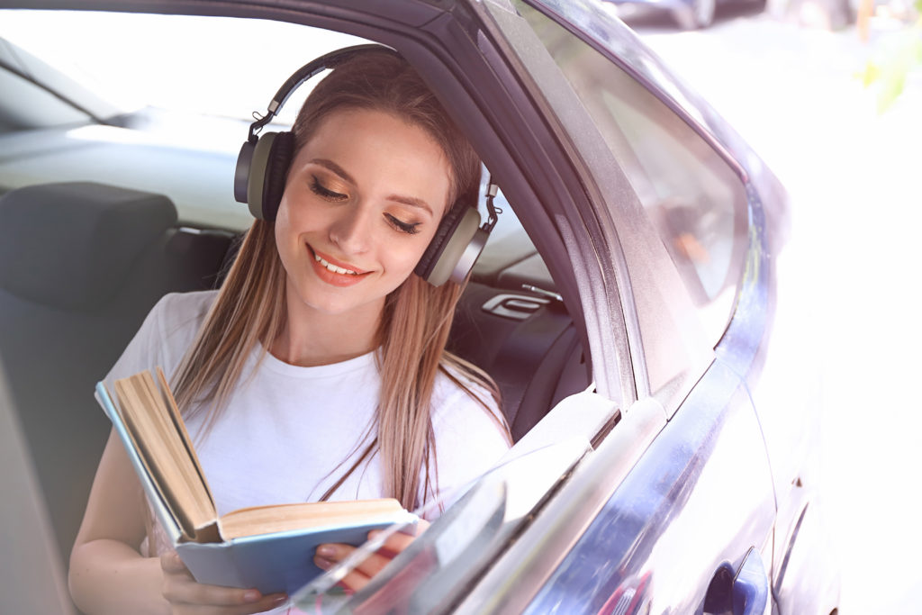 Beautiful young woman listening to music while reading book in car; 