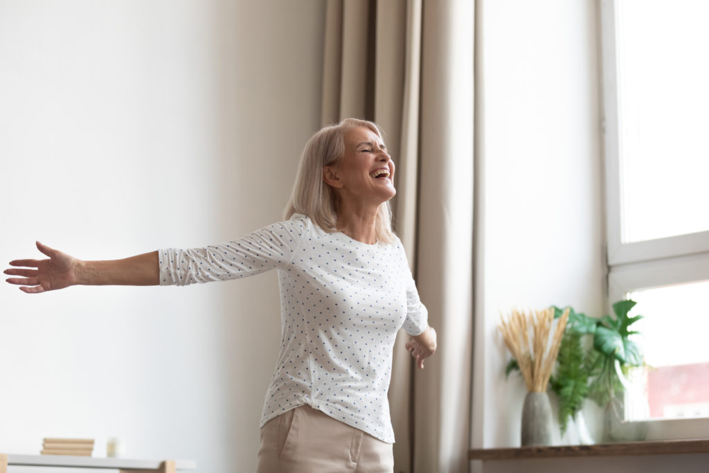 Laughing carefree middle-aged woman standing in living room stretched hands closed eyes breathing fresh air feels happy healthy, starts new day positive mood and thoughts, dancing enjoy life concept; 