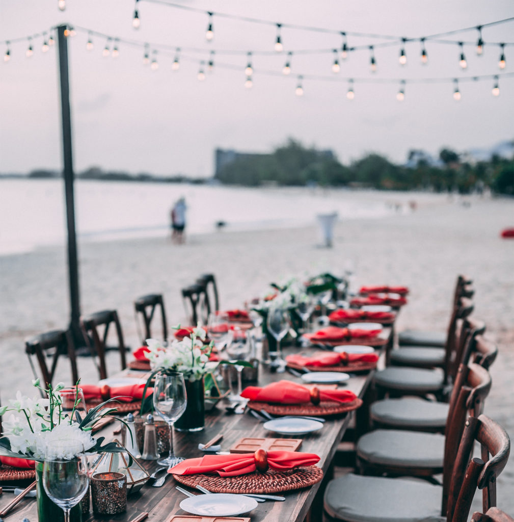 Long table beside beach., lights strung overhed, red napkins and plants on table