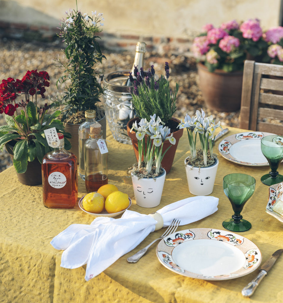 Table outdoors, yellow cloth, green wine glasses, pot plants on table, 2 with smiley faces drawn on the pots