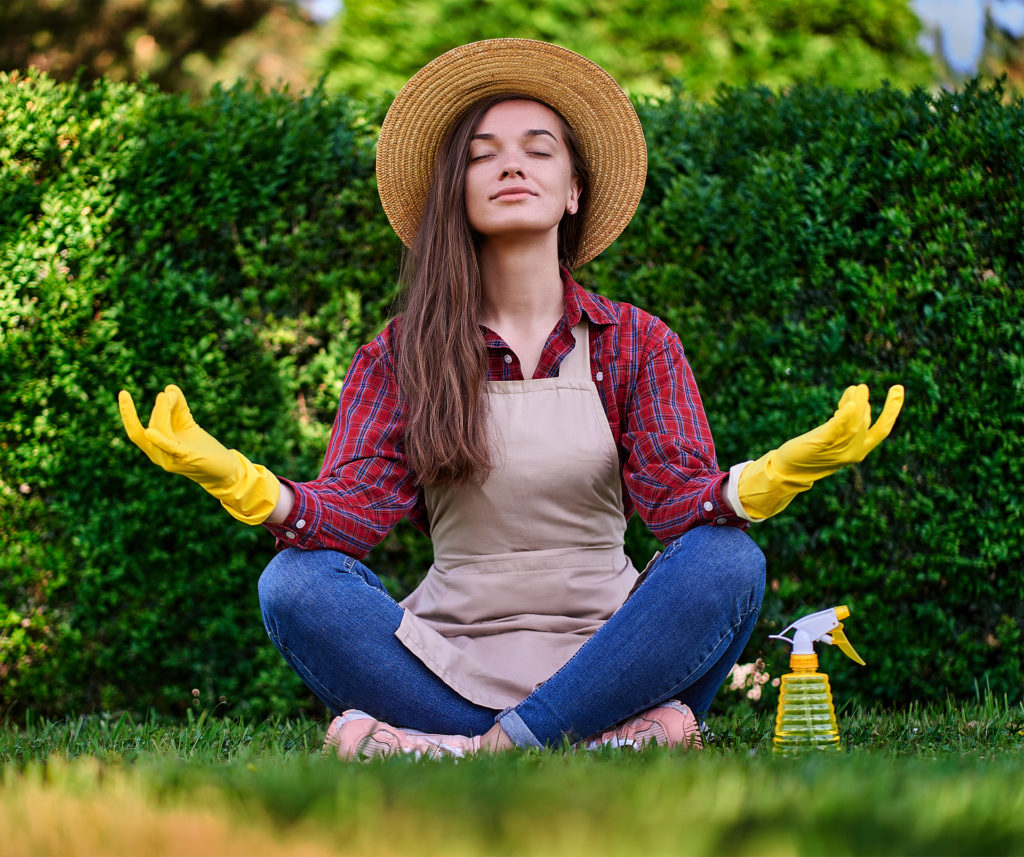 Calm woman gardener in a straw hat, apron and yellow rubber gloves in lotus pose meditates and relaxes on grass in green home garden. Mental health and enjoying of gardening