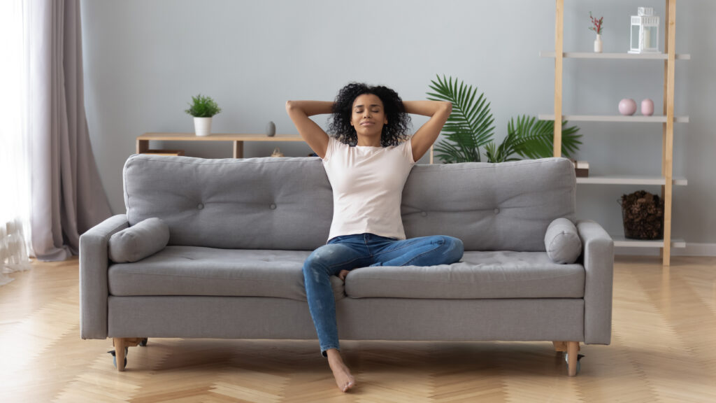 Young calm black woman relaxing sit on comfortable sofa in modern living room, lazy happy african woman girl resting on couch breathing fresh air enjoy peace of mind no stress free on couch at home; 
