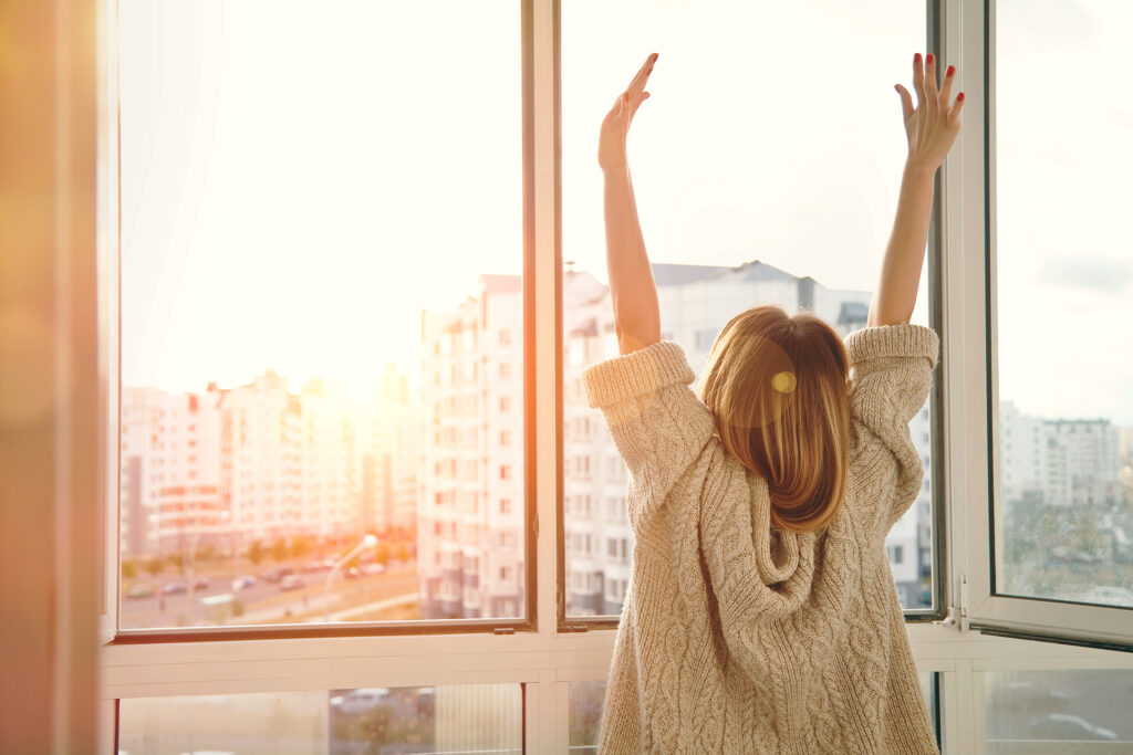 Woman near window raising hands facing the sunrise at morning