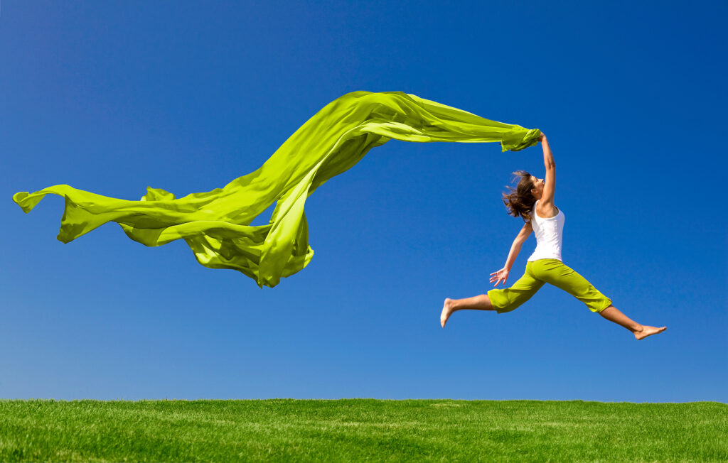 Beautiful young woman jumping on a green meadow with a colored tissue;