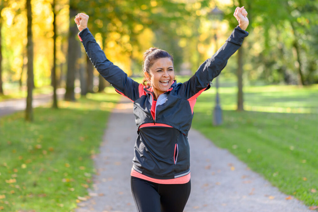 Happy fit middle aged woman cheering and celebrating as she walks along a rural lane through a leafy green park after working out jogging