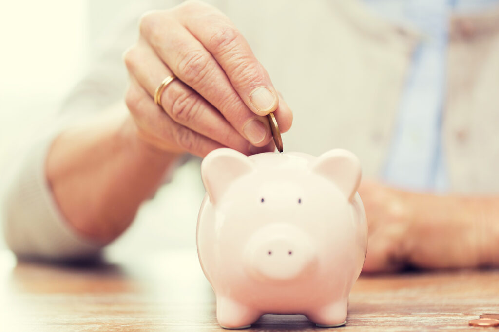 savings, money, annuity insurance, retirement and people concept - close up of senior woman hand putting coin into piggy bank; 