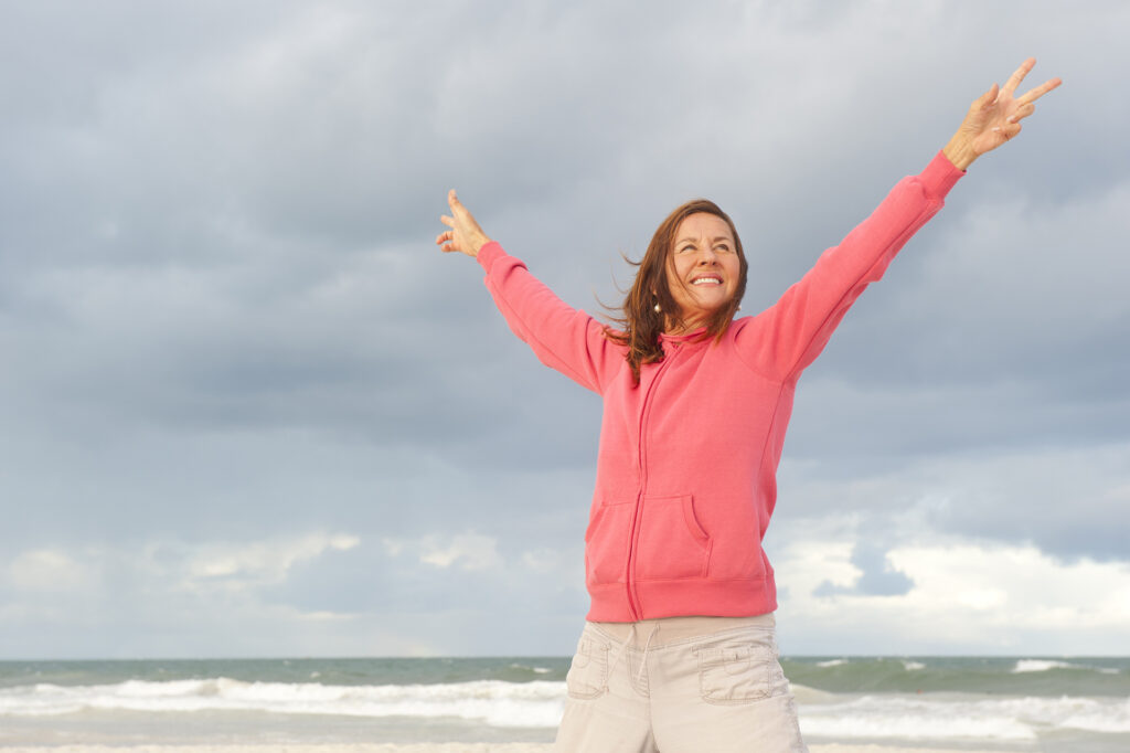 Portrait of positive and happy mature woman at the beach, wearing pink sweater, isolated with ocean and storm clouds as background and copy space.; 